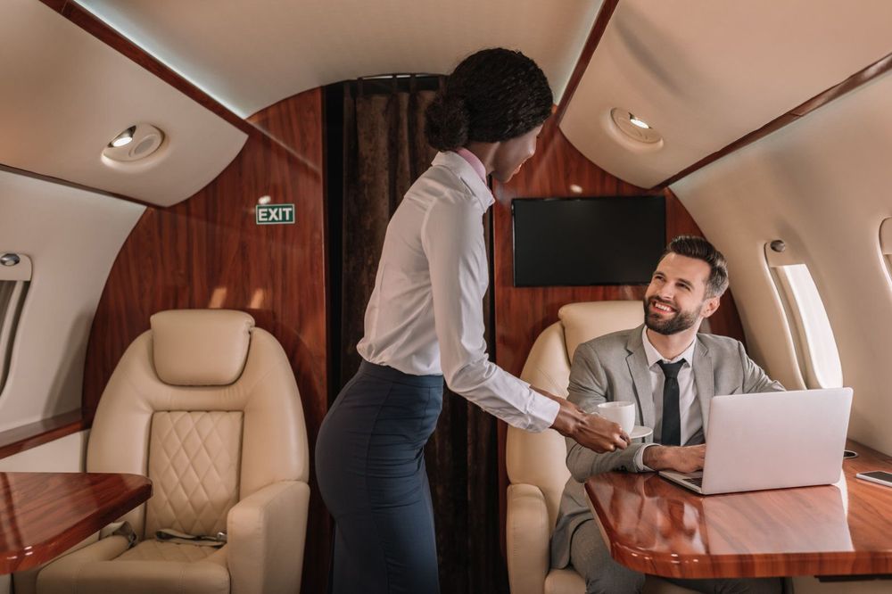 Beautiful young African American flight attendant dressed professionally hands a cup of coffee to a man in a suit with a smile and a laptop on a Private Jet to Miami.