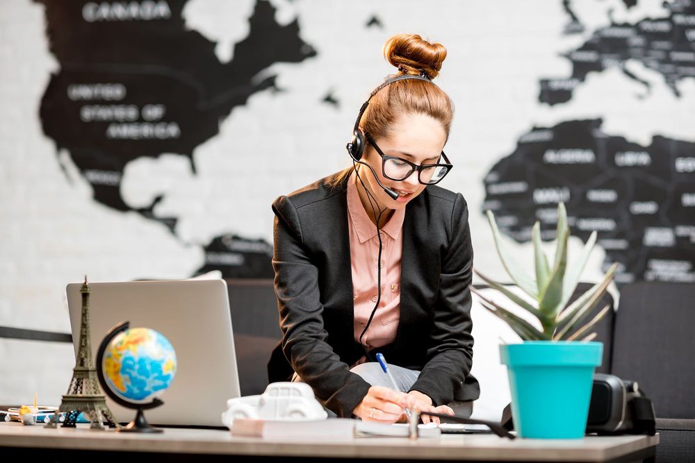A professionally dressed with hair bun, female travel concierge with Luxuri assisting a travel on the phone and via computer making sure they have support they need.